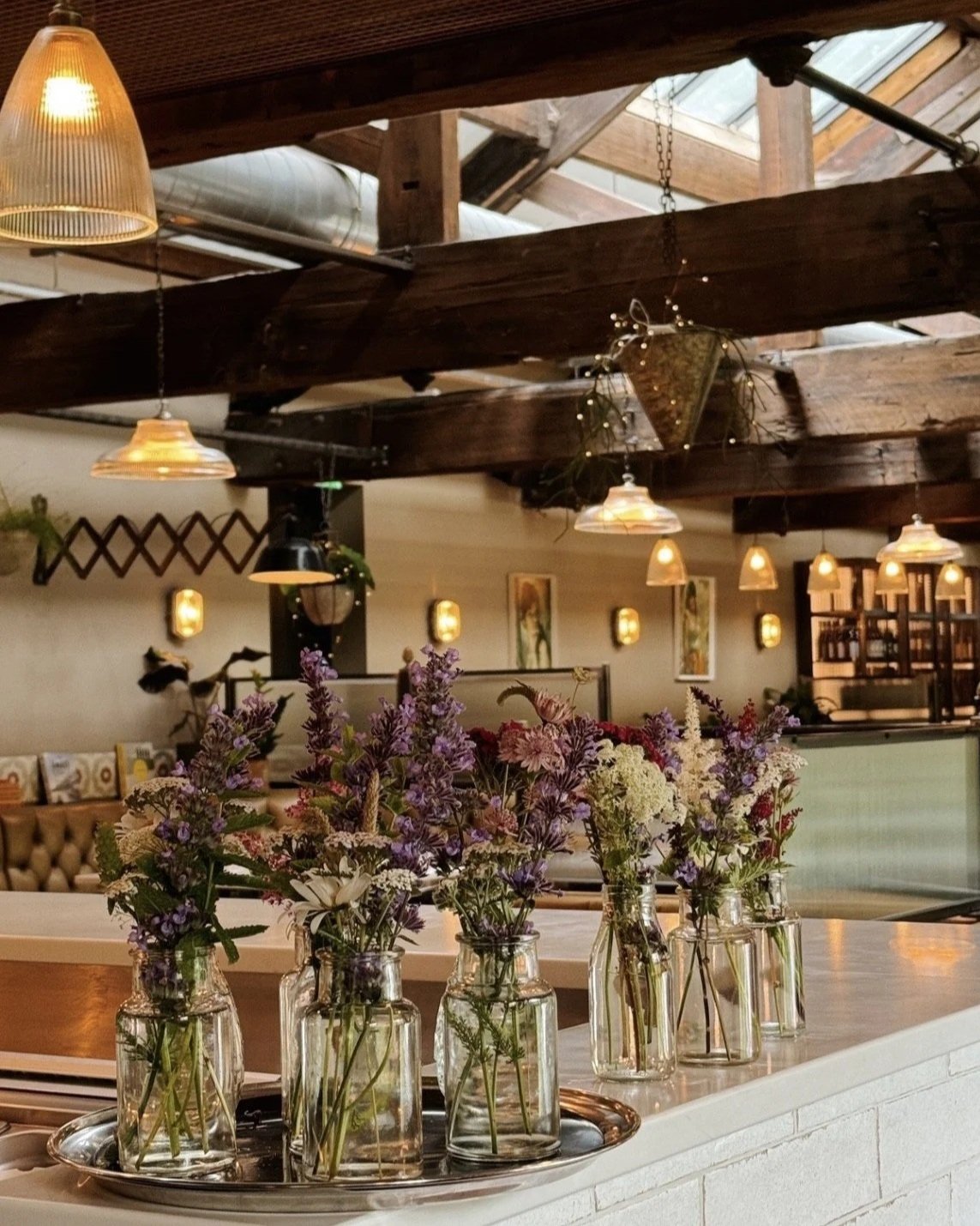 Numerous floral arrangements waiting to be set in a restaurant covered with high rustic wooden beams with rows of unique dangling lights.