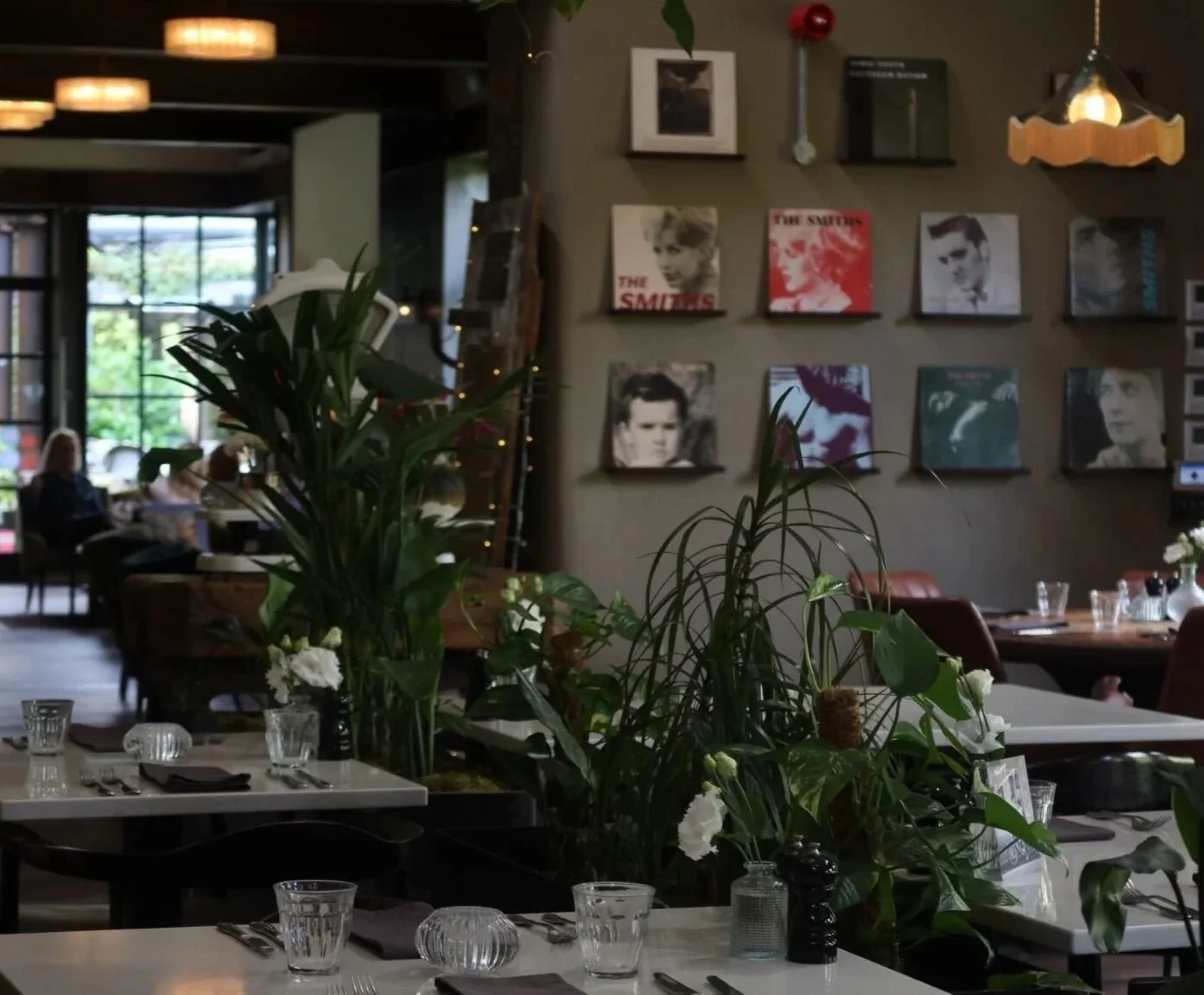 A cozy restaurant interior with round and rectangular tables set with placemats, glasses, and small flower arrangements. The walls feature framed photos and records, with ample greenery from potted plants near the windows. Warm lighting creates an in