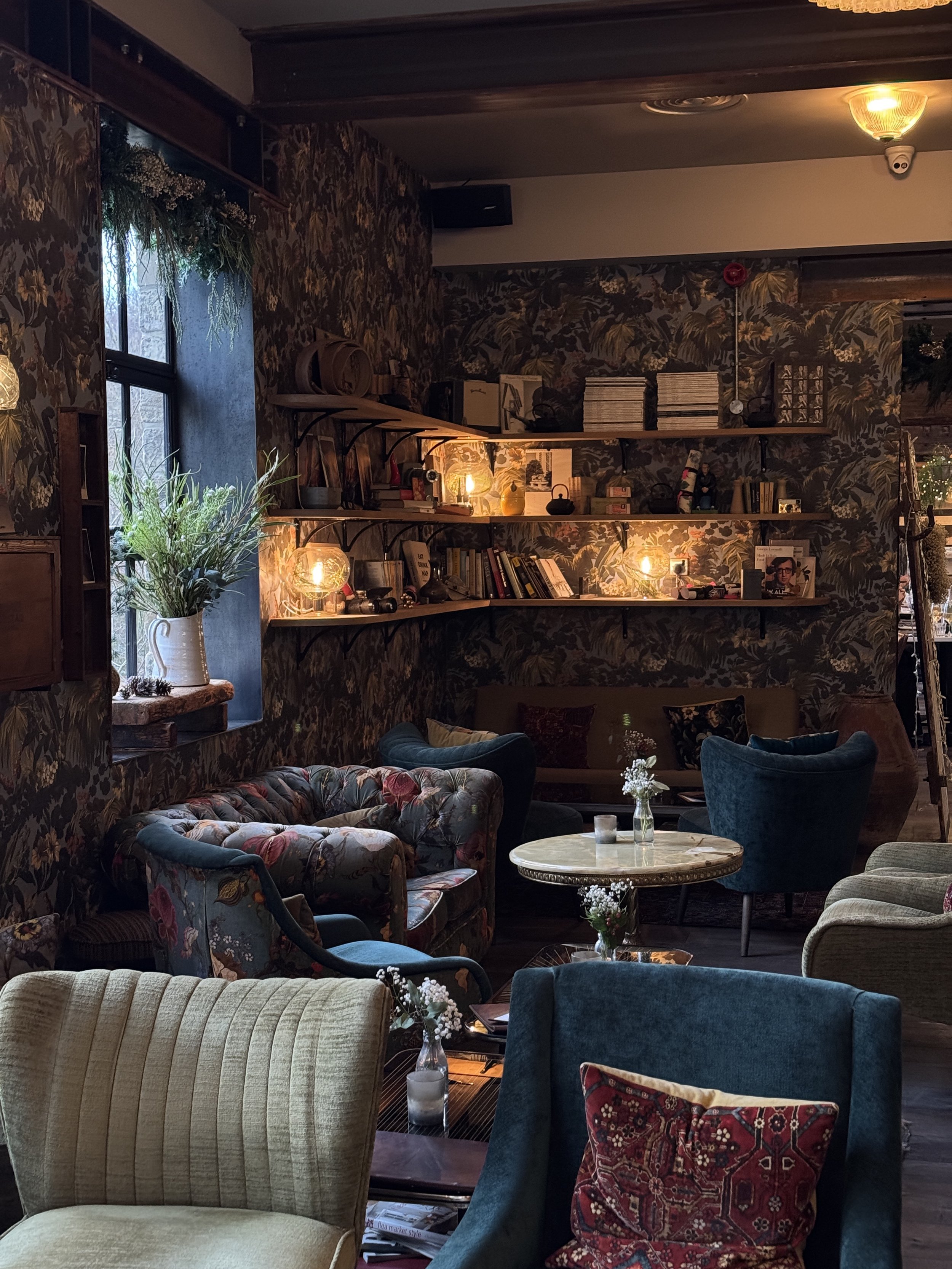 Cozy, vintage-style living room with floral wallpaper, plush armchairs, a sofa, and shelves with books and warm lighting.
