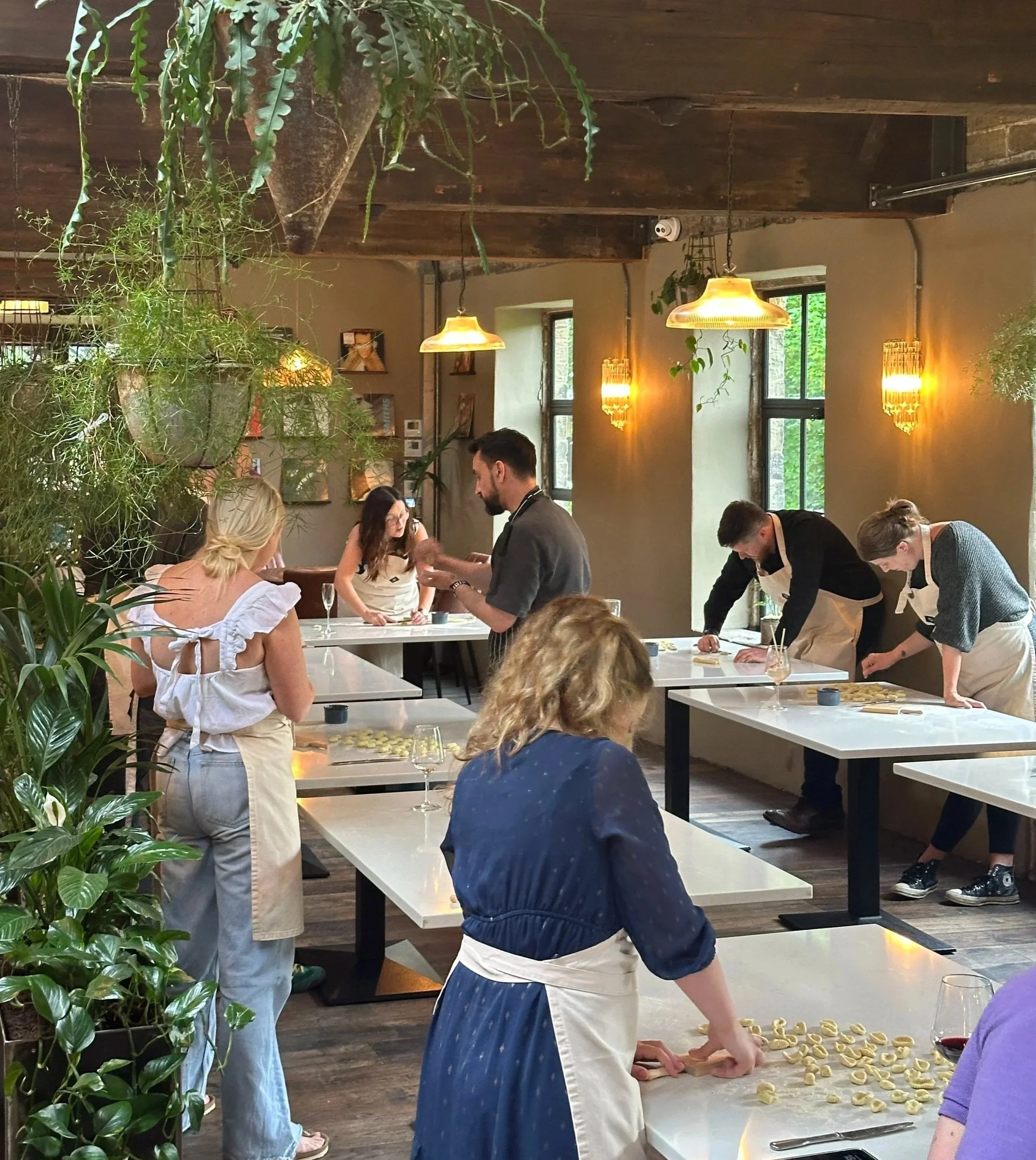 Wide shot of a pasta making class at Devour in Holmfirth, with guests of all ages and skill levels learning the Italian way to make gnocchi alongside Head Chef Carl, wine, prosecco and Aperol spritzes on the tables.