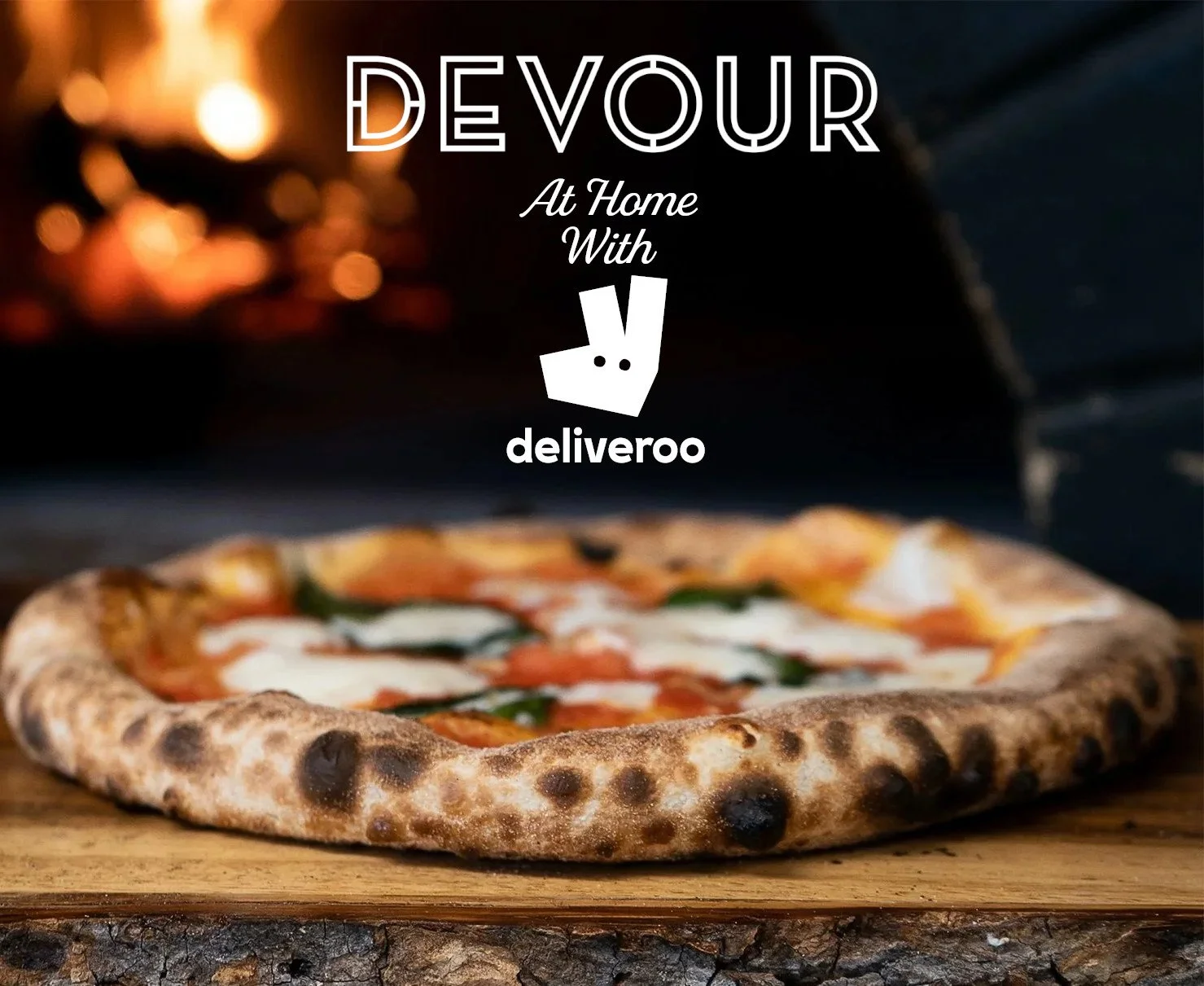 A wood-fired pizza on a log cutting board in front of a glowing pizza oven, overlaid with text reading “Devour at Home with Deliveroo” alongside Devour and Deliveroo logos.