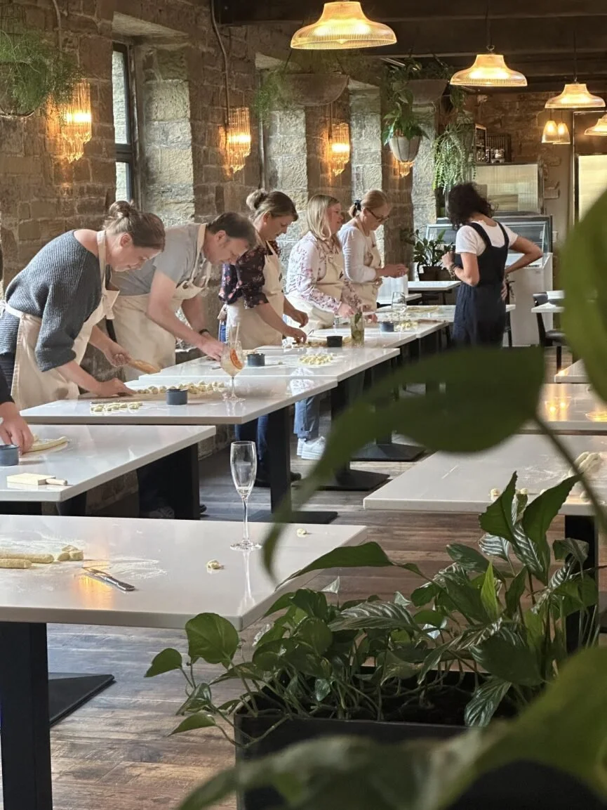 People participating in a Pasta and Pizza Cooking class or workshop, working with dough and tools at tables in a well-lit room, while being assisted by a teacher.