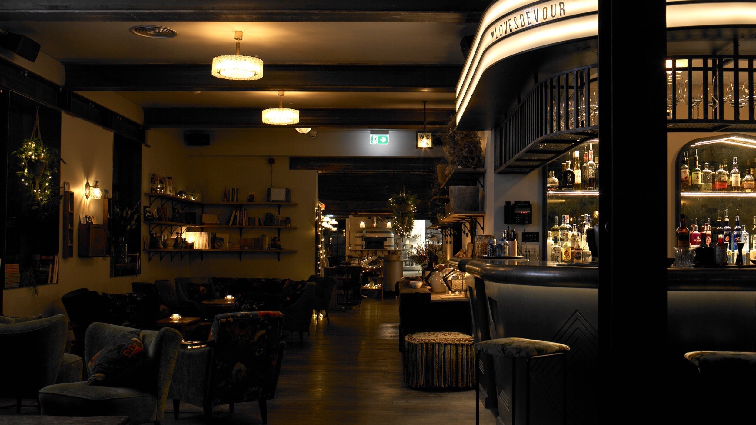 Dimly lit interior of a bar and lounge with shelves of liquor bottles behind the counter, cozy seating, and ambient lighting.