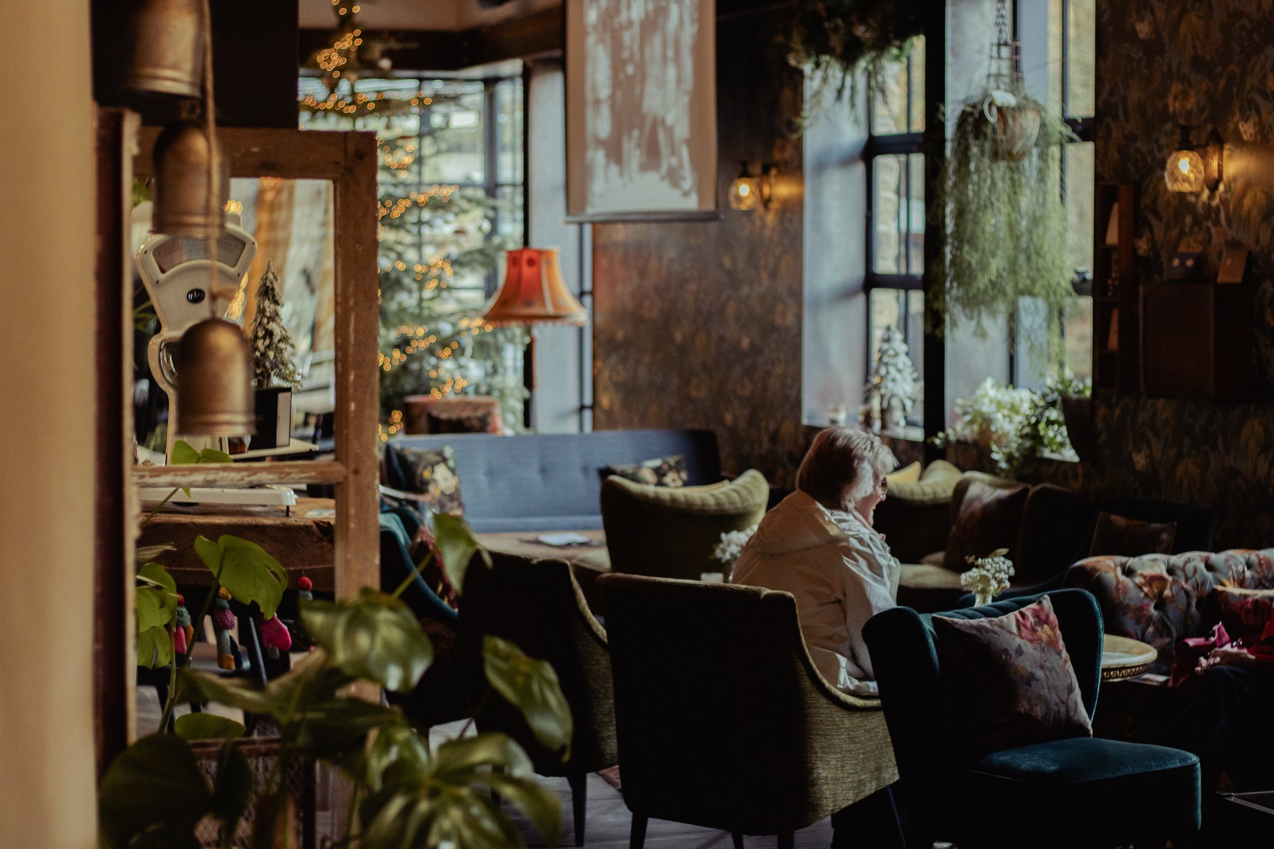 Cozy cafe with Christmas decorations, including a Christmas tree with lights, and a woman sitting alone at a table near large windows.