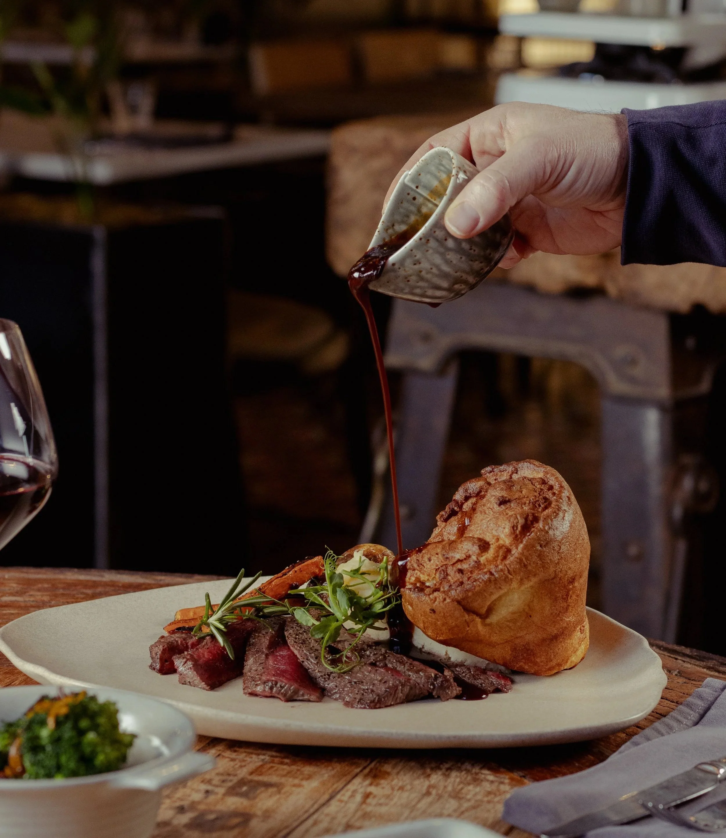 Plate of roasted meat with mashed potatoes, fried vegetables, and mushrooms on a rustic wooden table in a cozy restaurant setting with soft lighting and a flower arrangement in the background.