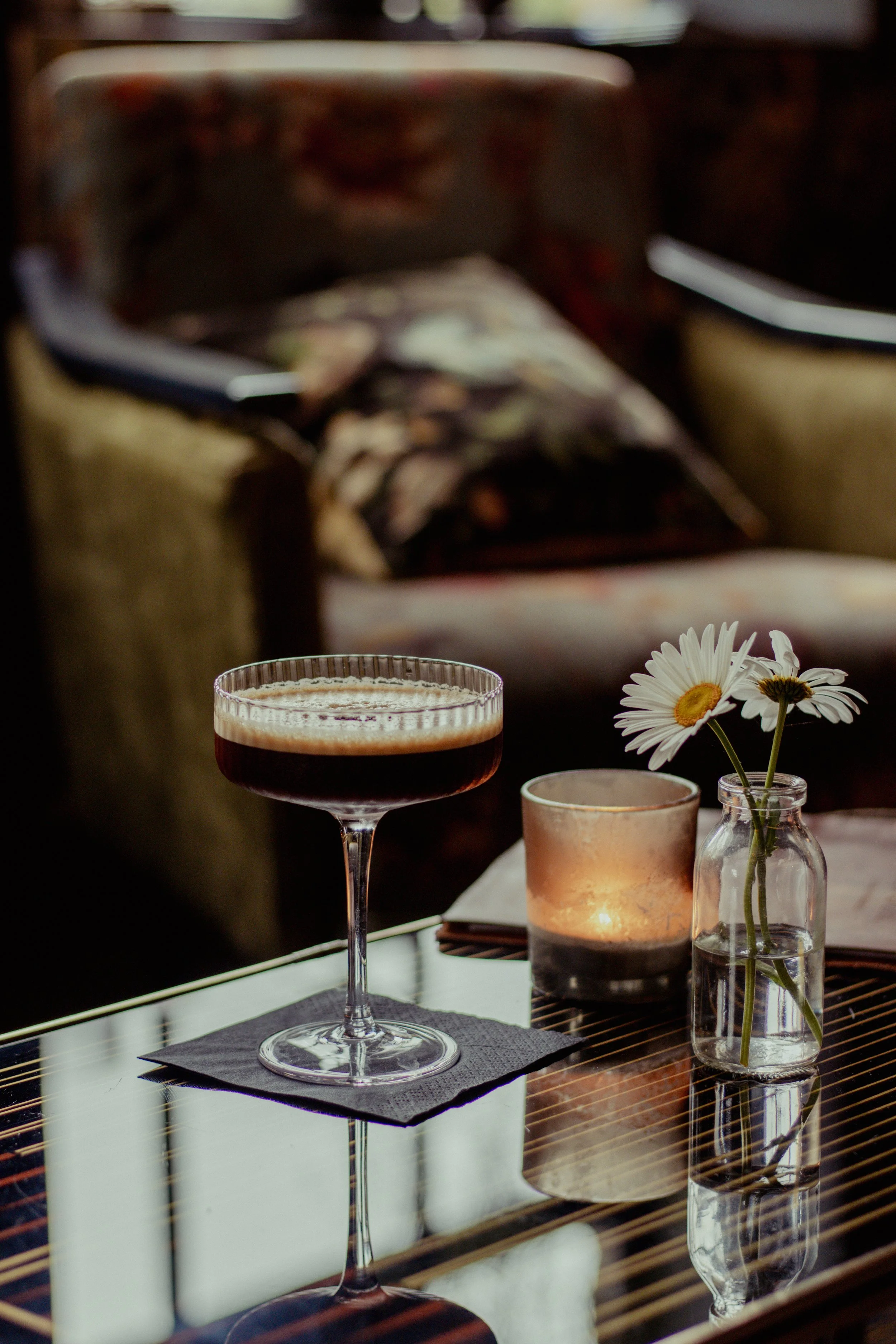 A cocktail glass with a dark beverage and frothy top, a lit candle in a glass holder, and a small vase with white daisies on a reflective table.