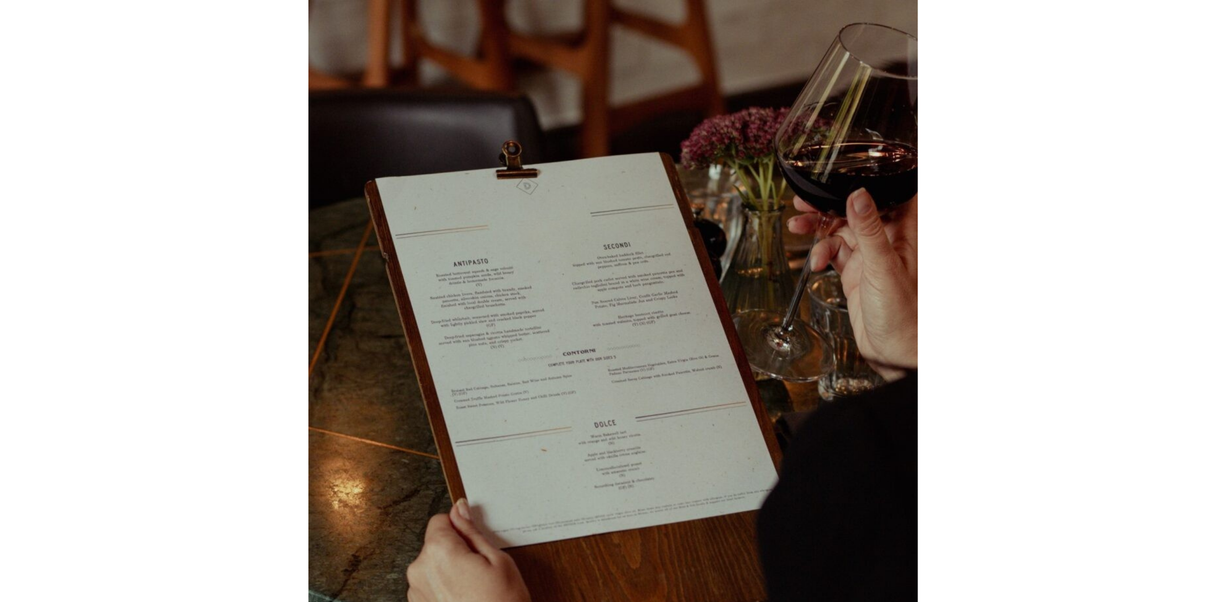 A woman holding a menu in one hand and a elegant glass of red wine in her other hand, she is sat on a marble green table with gold accents with purple flowers in a vase adding colour to the scene while in the distance the background is blurred