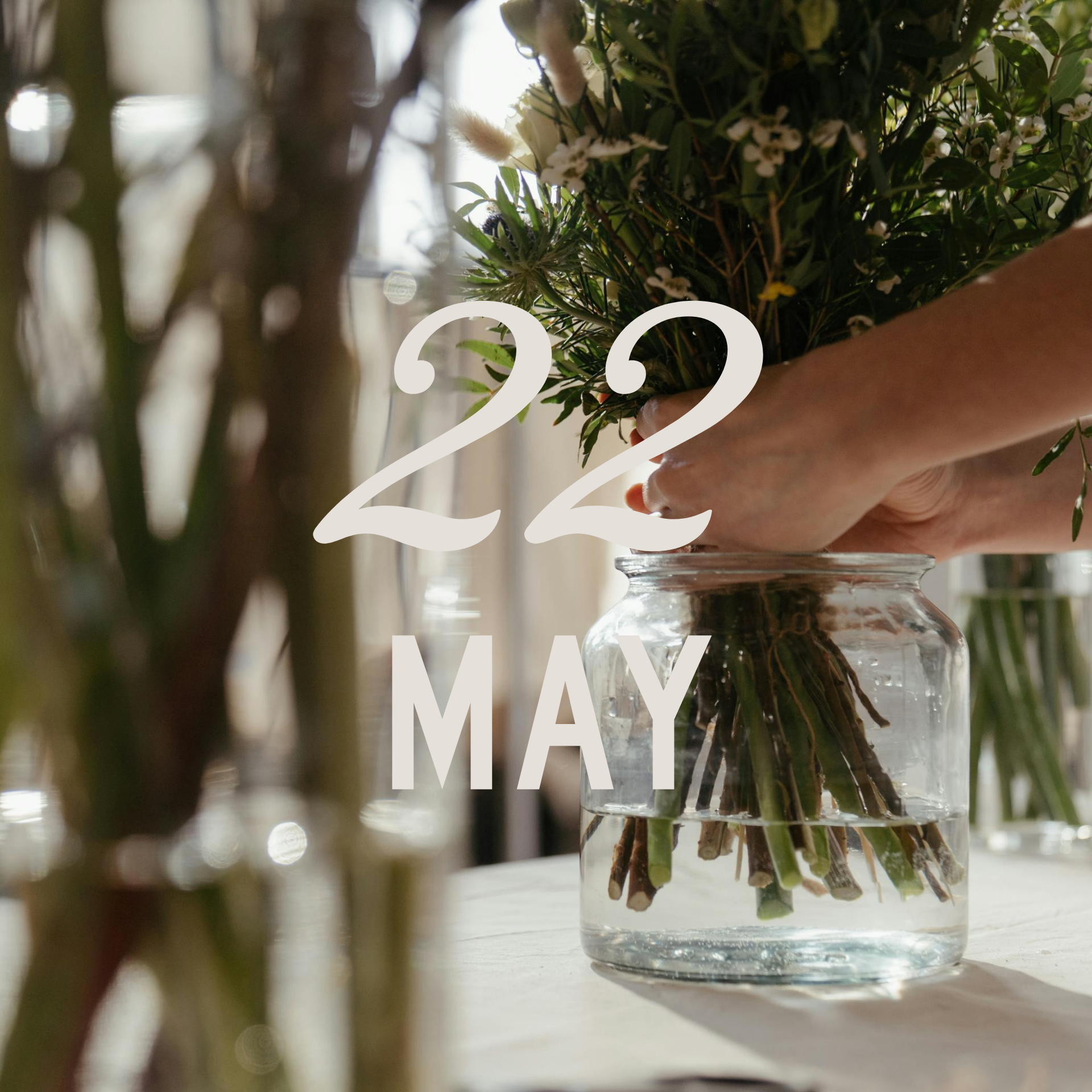 Close-up of a glass jar with water holding a bouquet of assorted flowers and green stems, with a blurred background.