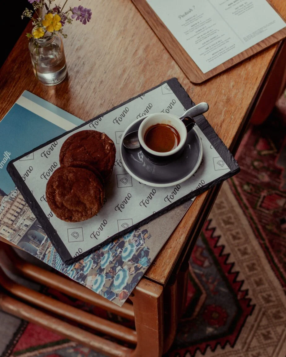 An espresso coffee sat beside two double chocolate cookies on a slate plate covered in Devour branding next to a flower pot and drinks menu. While, sat on top of an elegant art-deco wooden table and cushioned by a cosy red patterned rug. 