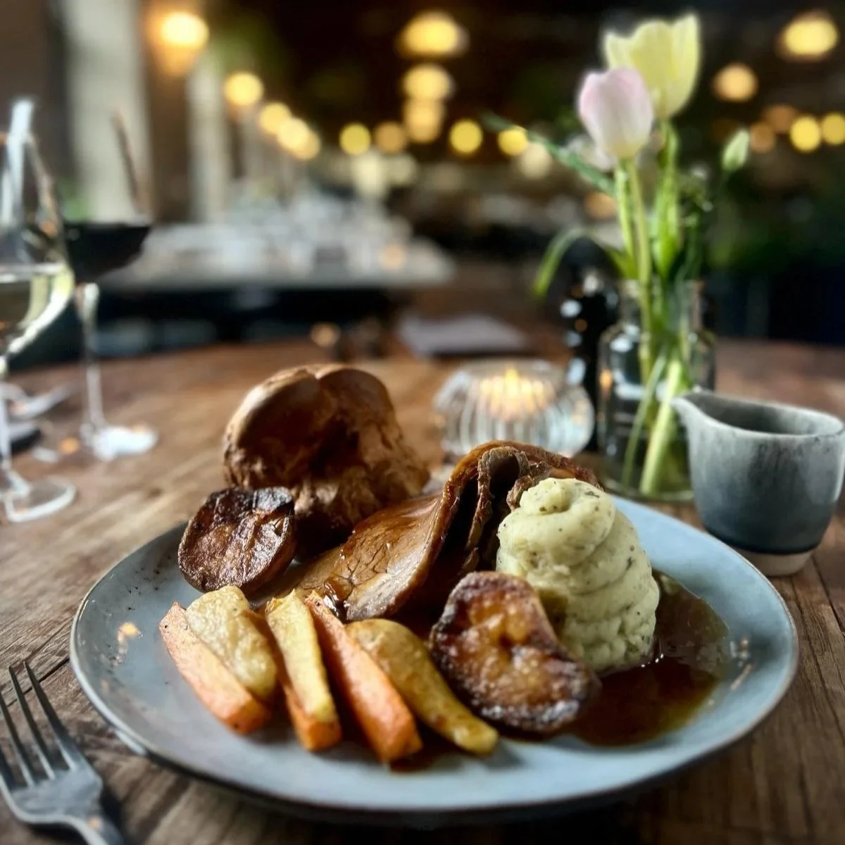 A Devour Sunday lunch featuring roast beef, mash, roast potatoes and carrots on a wooden table, surrounded by red and white wine glasses, gravy boat and flowers, with Devour’s cosy, softly lit restaurant blurred in the background.