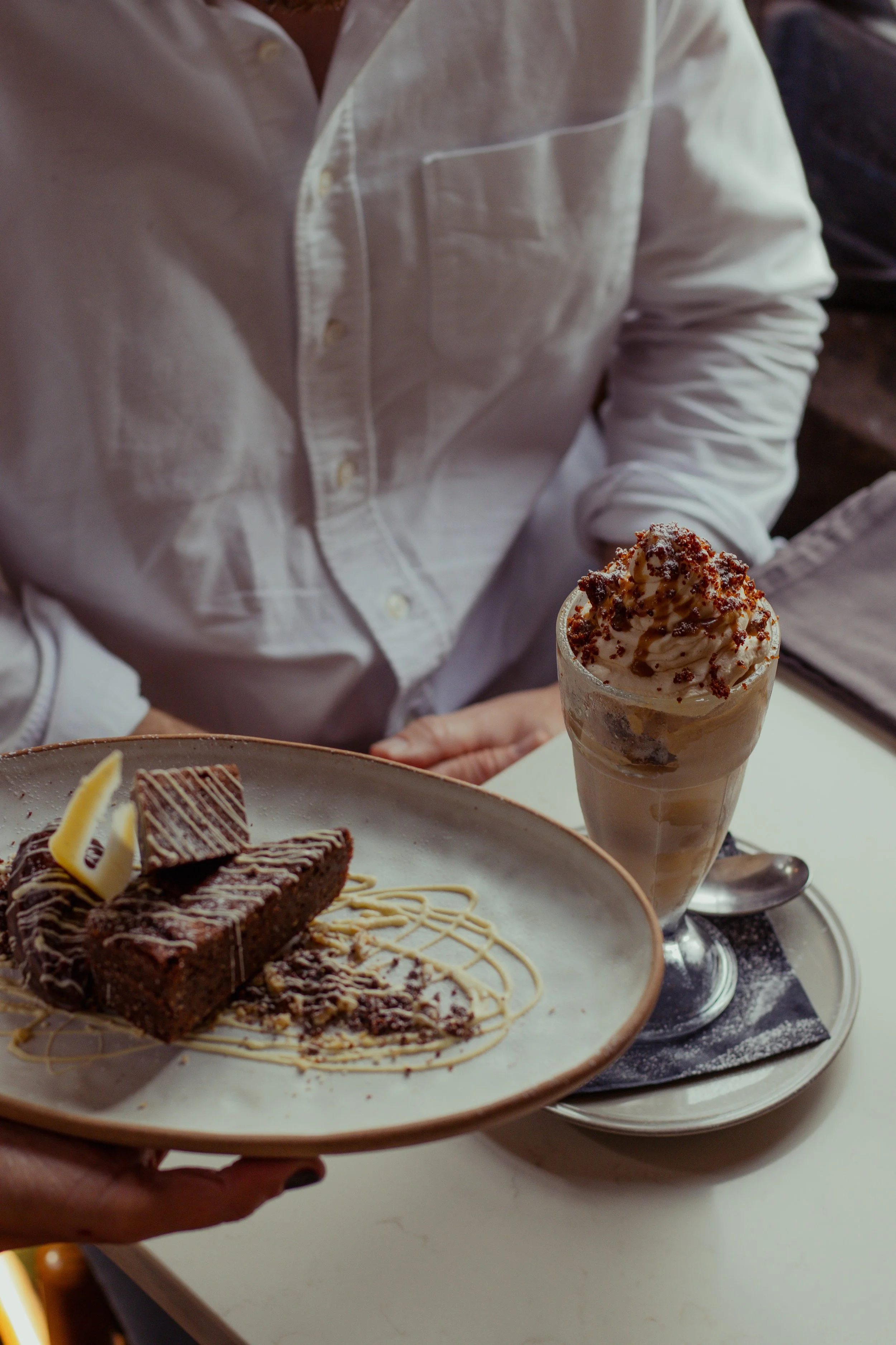 A person holding a plate with a slice of chocolate cake and a lemon wedge, with a dessert drink topped with whipped cream and caramel sauce nearby.