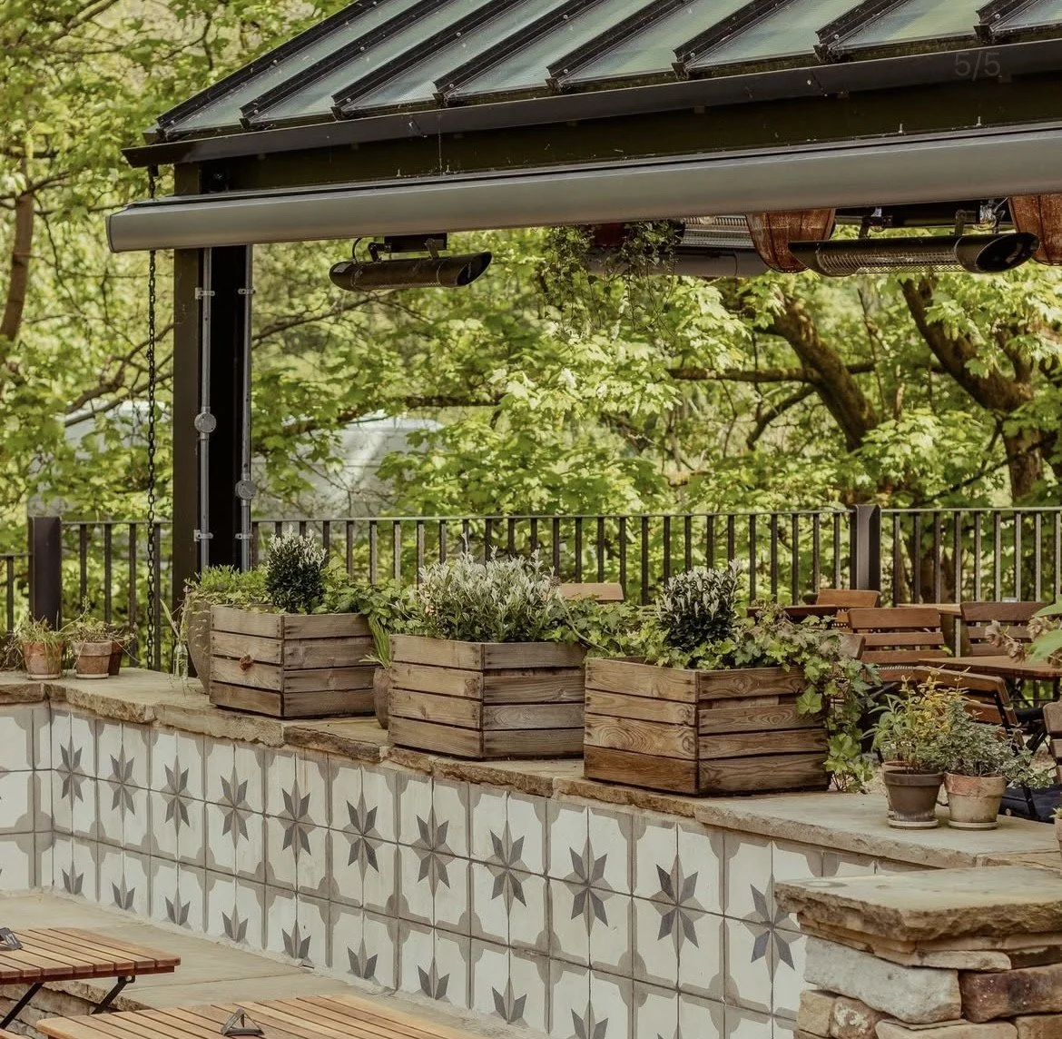 A beautifully green outdoor eating space. Wooden tables. Cosy benches. Beautifully tiled walls and wooden box plant pots, create a cosy, outdoor space enclosed by nature. Overhead is a greenhouse roof fit with heaters. Trees and nature set the scene.