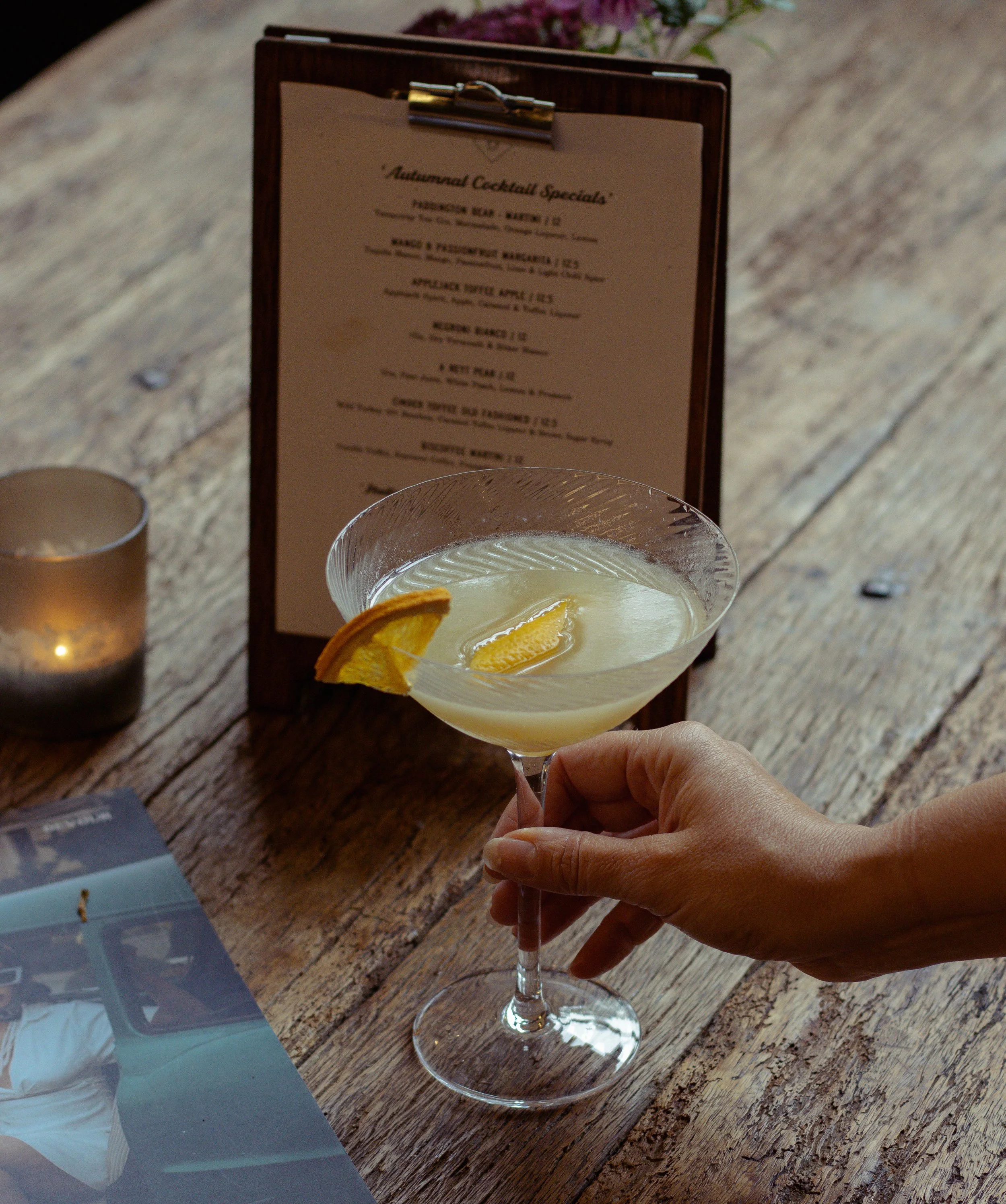 A cocktail glass with a orange liquid and garnished with an orange peel and slice, placed on a rustic wooden table, next to a menu and lit by candle-light.