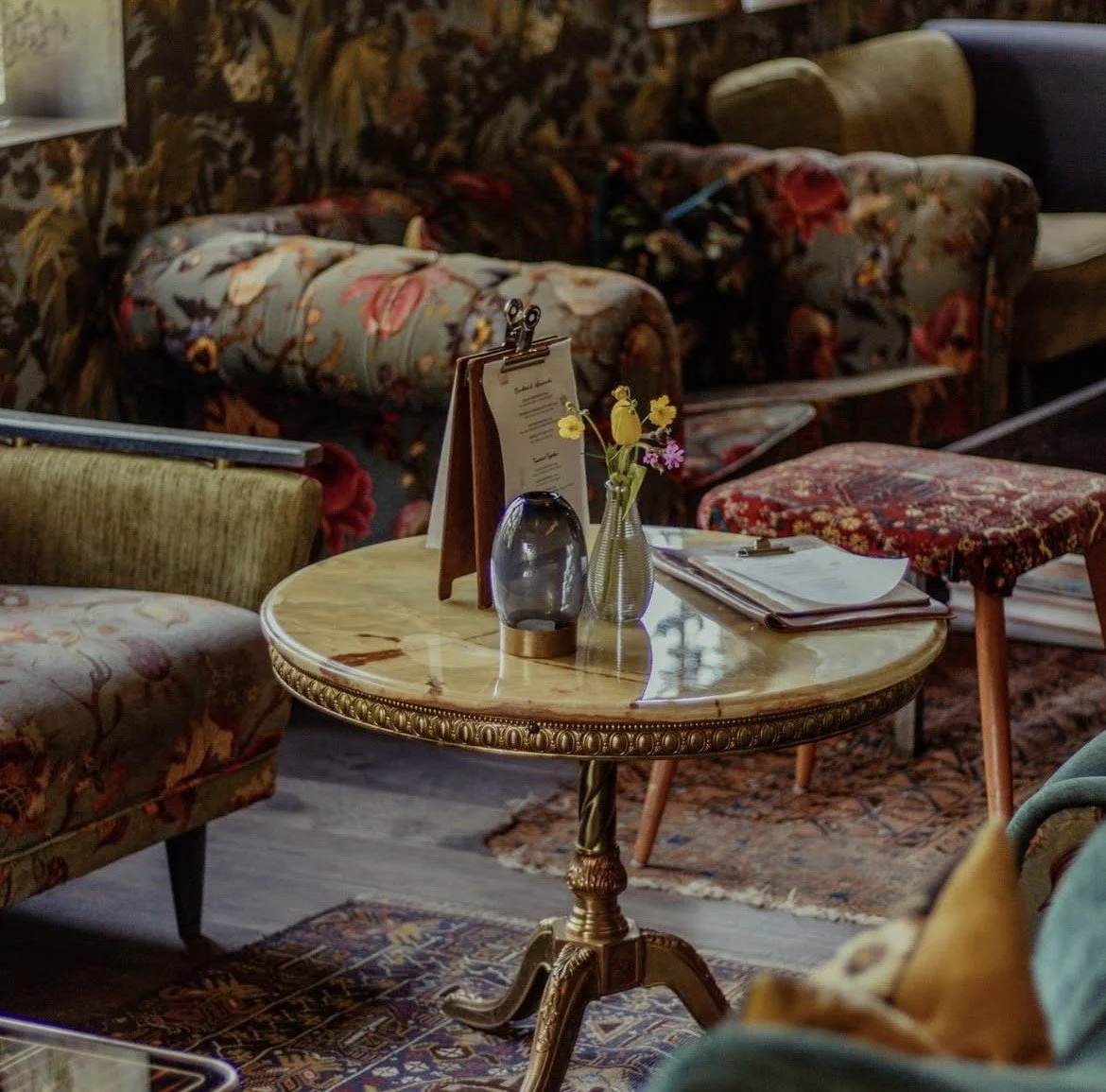 Cozy, vintage-style bar area with floral wallpaper, plush armchairs, a sofa, and shelves surrounding an antique table with a menu, candle and colourful flowers on top.