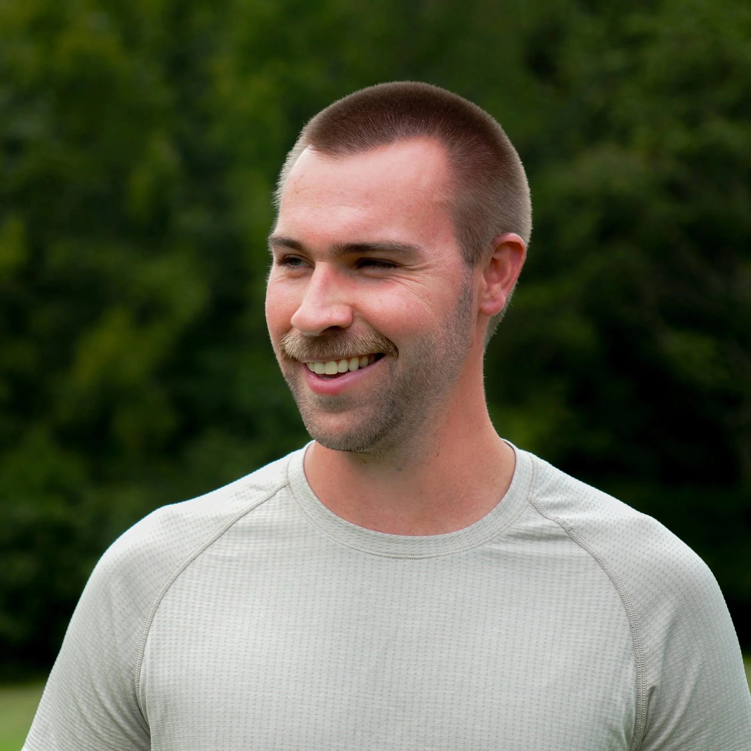 A smiling man with a trimmed beard and short hair wearing a light gray athletic shirt outdoors with green trees in the background.