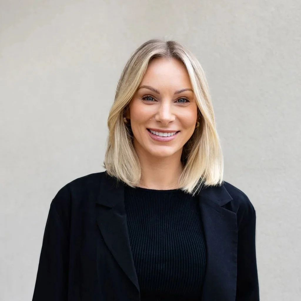 A woman with blonde hair smiling and leaning against a wall, wearing a white top and earrings.