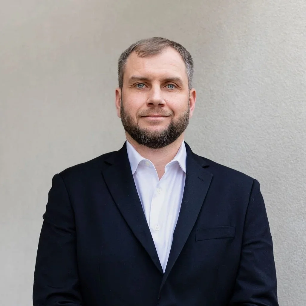 A man with light brown hair and beard, wearing a dark suit, white shirt, and red tie, standing against a plain beige background.