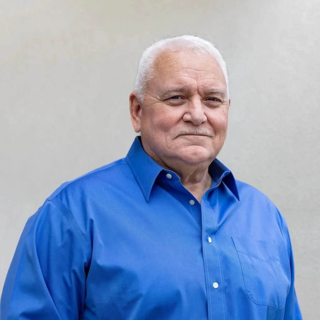 Portrait of an older man with white hair and a mustache wearing a light blue collared shirt, standing against a textured gray wall.