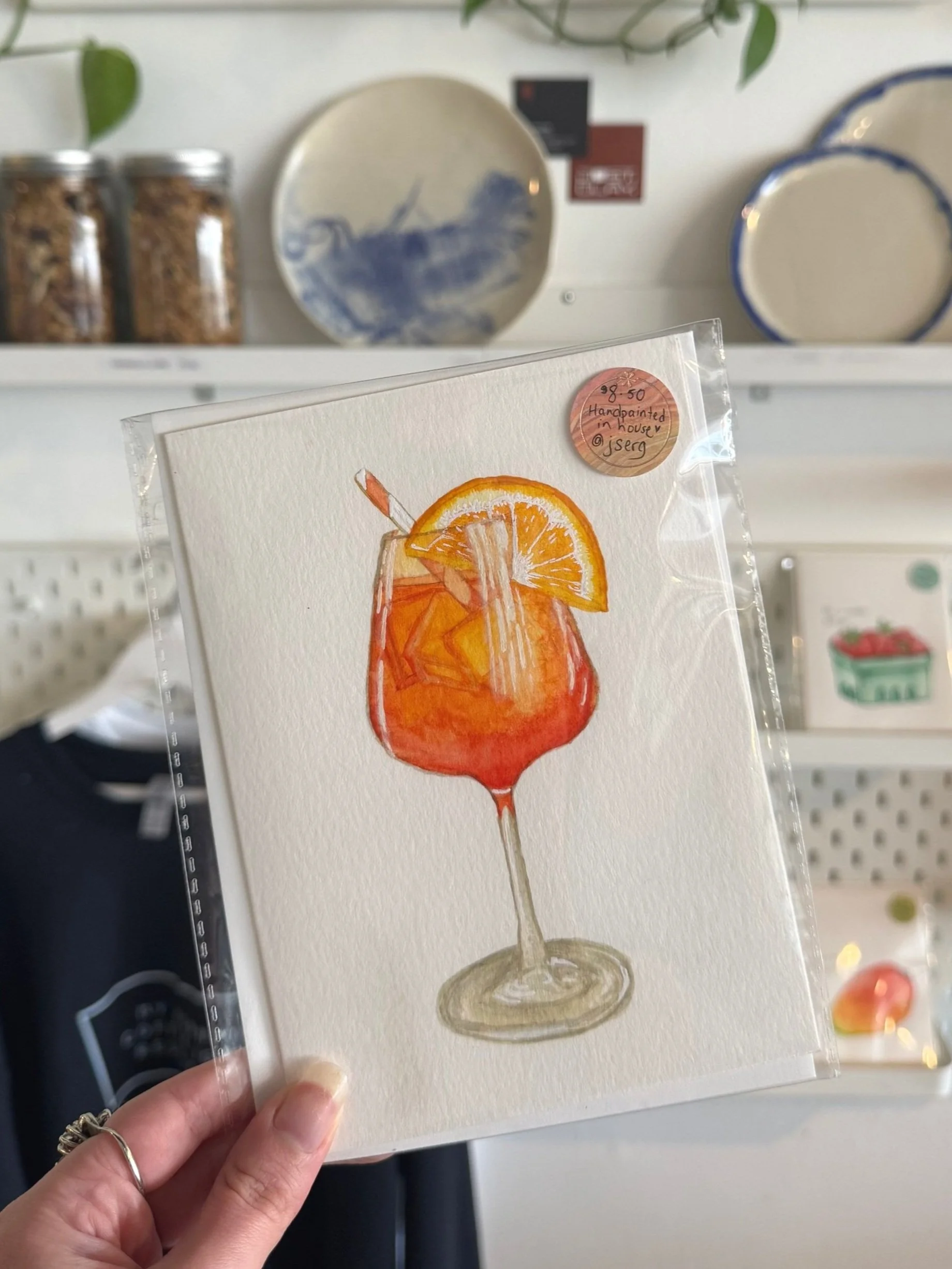 Watercolor painting of a cocktail glass filled with Aperol Spritz garnished with orange slices and a straw, held in front of ceramic plates and jars on a shelf.