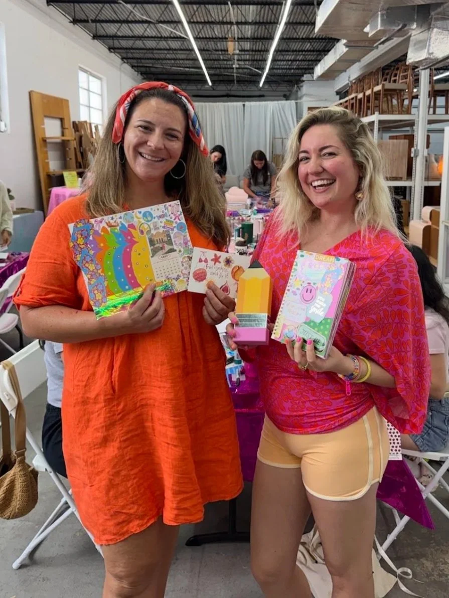 Two women smiling and holding their junk journal creations at an Art Club FTL workshop at Rehaus, a vintage furniture store in Oakland Park Florida. Some of the furniture as well as other Art Club guests are visible in the background.