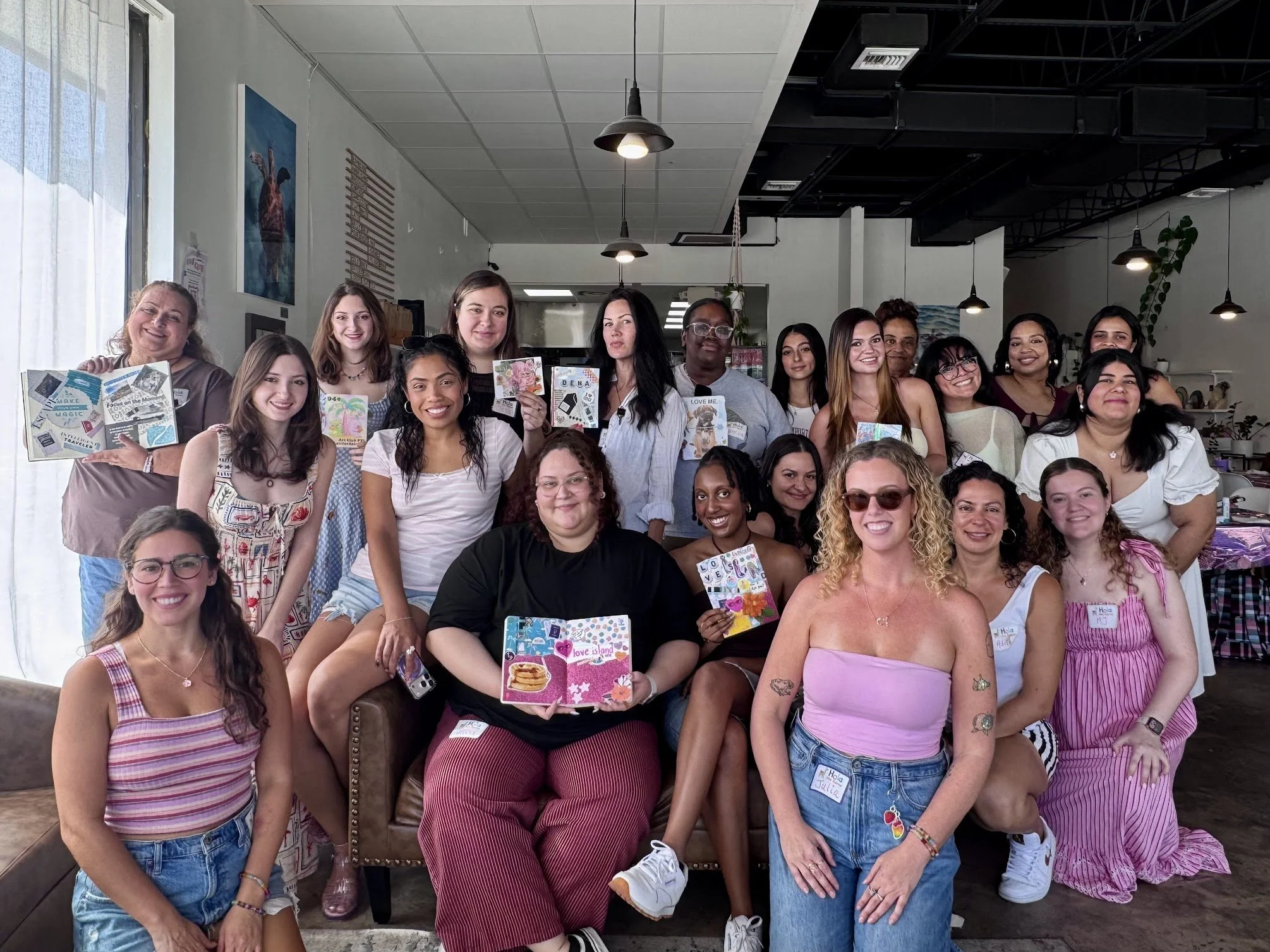 Group of women smiling and holding colorful, decorated notebooks or journals in a bright, modern indoor space with industrial lighting.