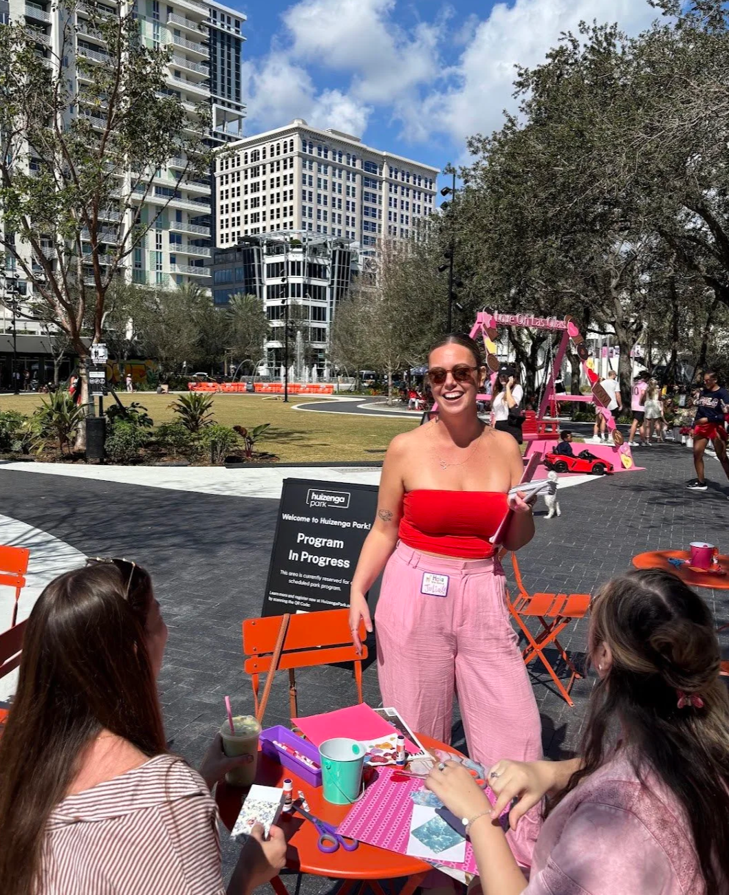 Three women sitting at a table outdoors in downtown Fort Lauderdale, junk journaling with Art Club, with one standing woman smiling at the camera. The scene is in in Huizenga Park with tall buildings and palm trees visible in the background.