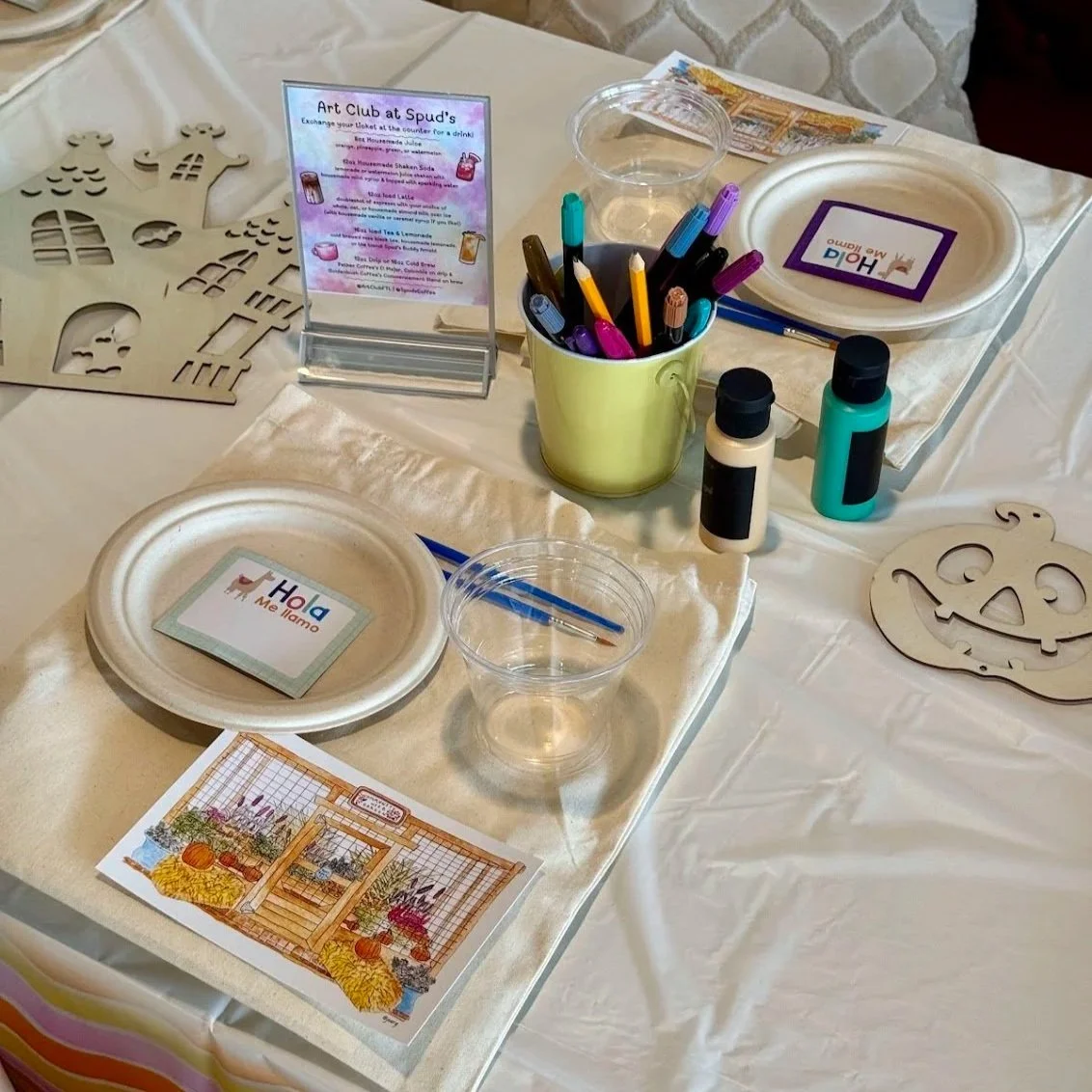 Craft table with wooden pumpkin and ghost cutouts, art supplies including markers, paint bottles, and a paintbrush, paper plates, a Halloween-themed sign, and a sign for an art club at Spud's.