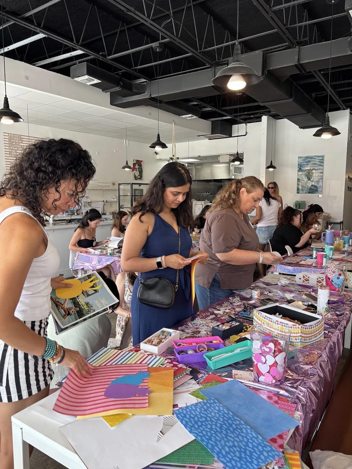 Women participating in the World's Largest Craft Club event by Art Club FTL at a table with colorful craft supplies at Spud's Coffee, a bright coffeeshop in Fort Lauderdale.