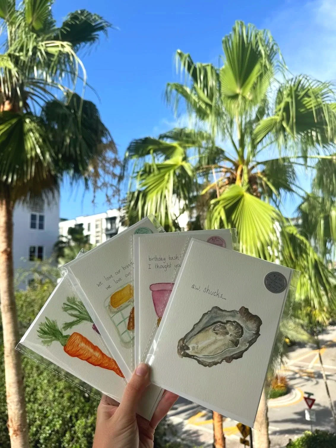 Hand holding four illustrated greeting cards featuring a carrot, a cake, an oyster, and a breadstick, with a tropical backdrop of palm trees and a blue sky.
