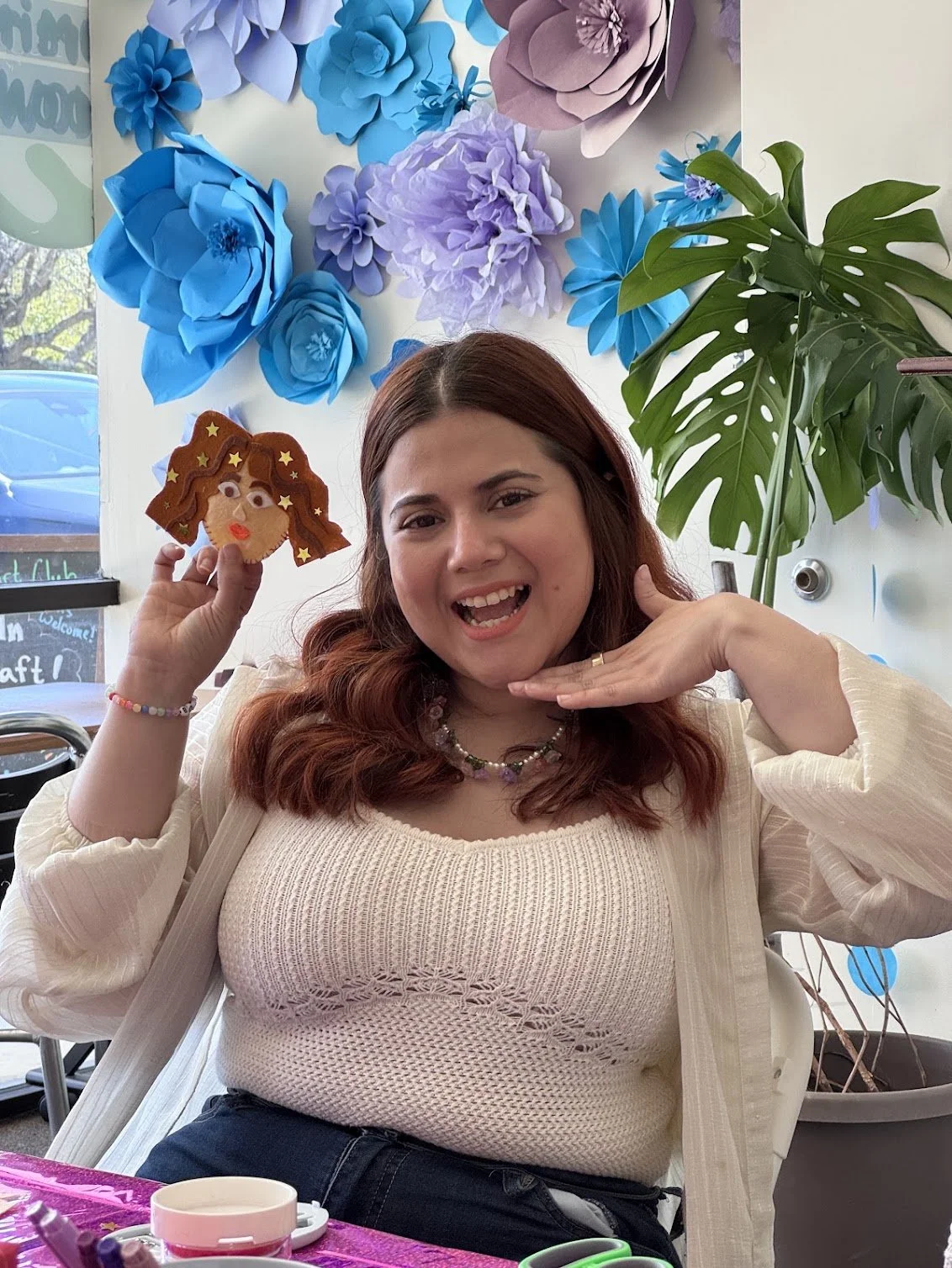 A woman with reddish hair smiling and posing for a photo while holding a mini felt ornament version of herself at at Art Club FTL workshop at Crafted Art Club in Coconut Creek. The background features a wall of handmade blue and purple paper flowers.