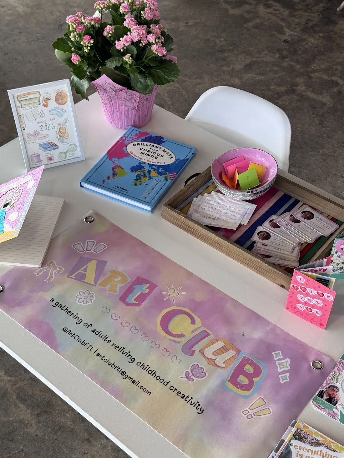 The table has a large pink and purple banner that reads 'ART CLUB' and 'a gathering of adults reliving childhood creativity.' There is a pink potted plant with pink flowers, a colorful book titled 'Brilliant Minds,' a small frame with illustrated notes, a bowl with colorful sticky notes, and various art and craft supplies, including stickers, cards, and paper.