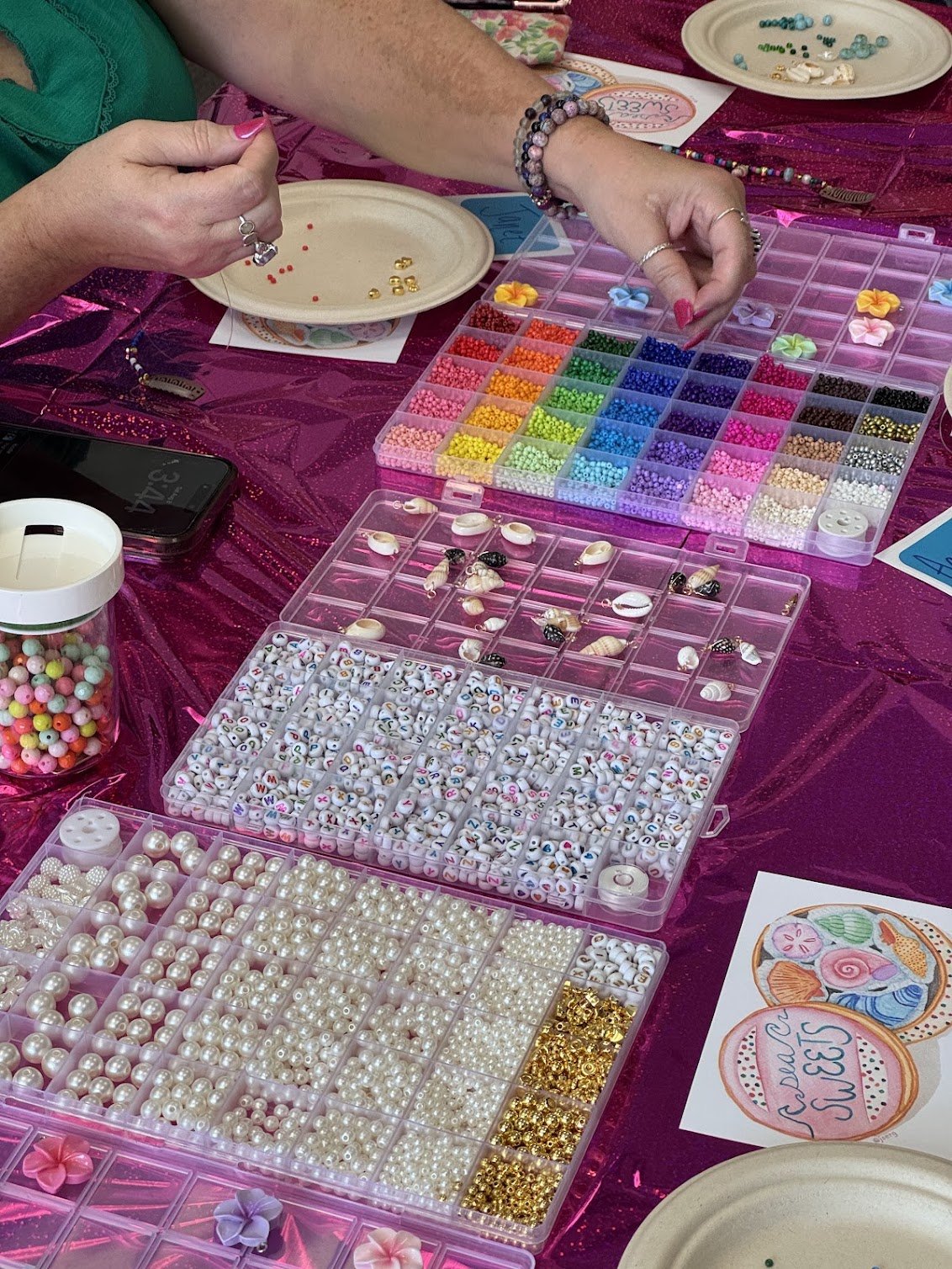 A person making a hair charm at an Art Club FTL workshop at Crafted Art Studio in Coconut Creek with various colorful beads, pearls, and charms organized in plastic containers on a pink tablecloth.