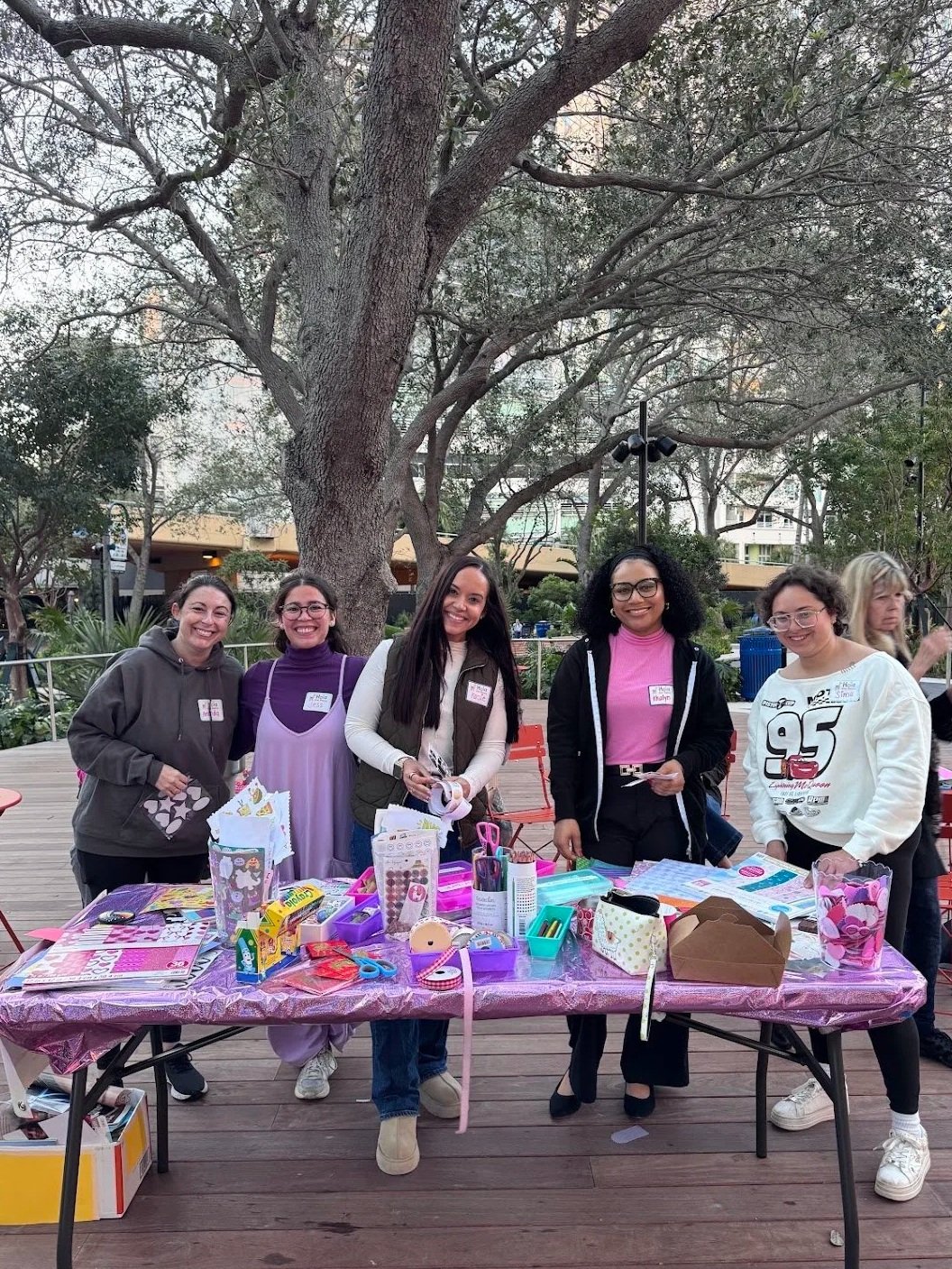 Group of five women standing behind a table with craft supplies  at an outdoor junk journal workshop by Art Club FTL in Huizenga Park in downtown Fort Lauderdale.. Large tree and greenery in the background.