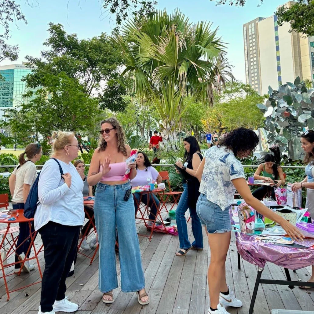 Women browsing shared arts and crafts supplies at an outdoor junk journal workshop by Art Club FTL in Huizenga Park in downtown Fort Lauderdale.