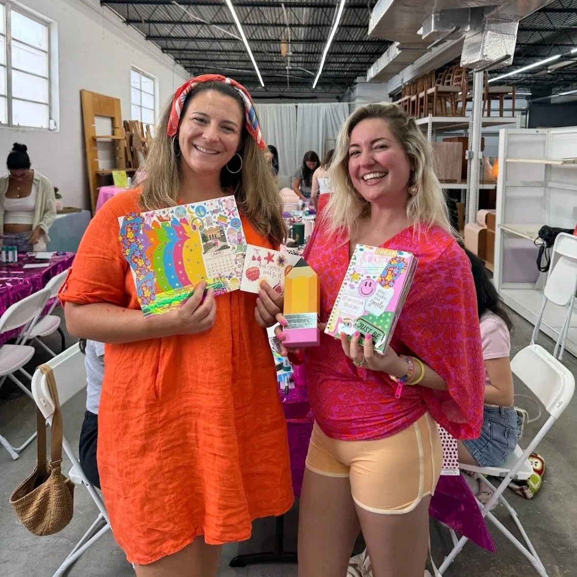 Two women smiling and holding their junk journal creations at an Art Club FTL workshop at Rehaus, a vintage furniture store in Oakland Park Florida. Some of the furniture as well as other Art Club guests are visible in the background.