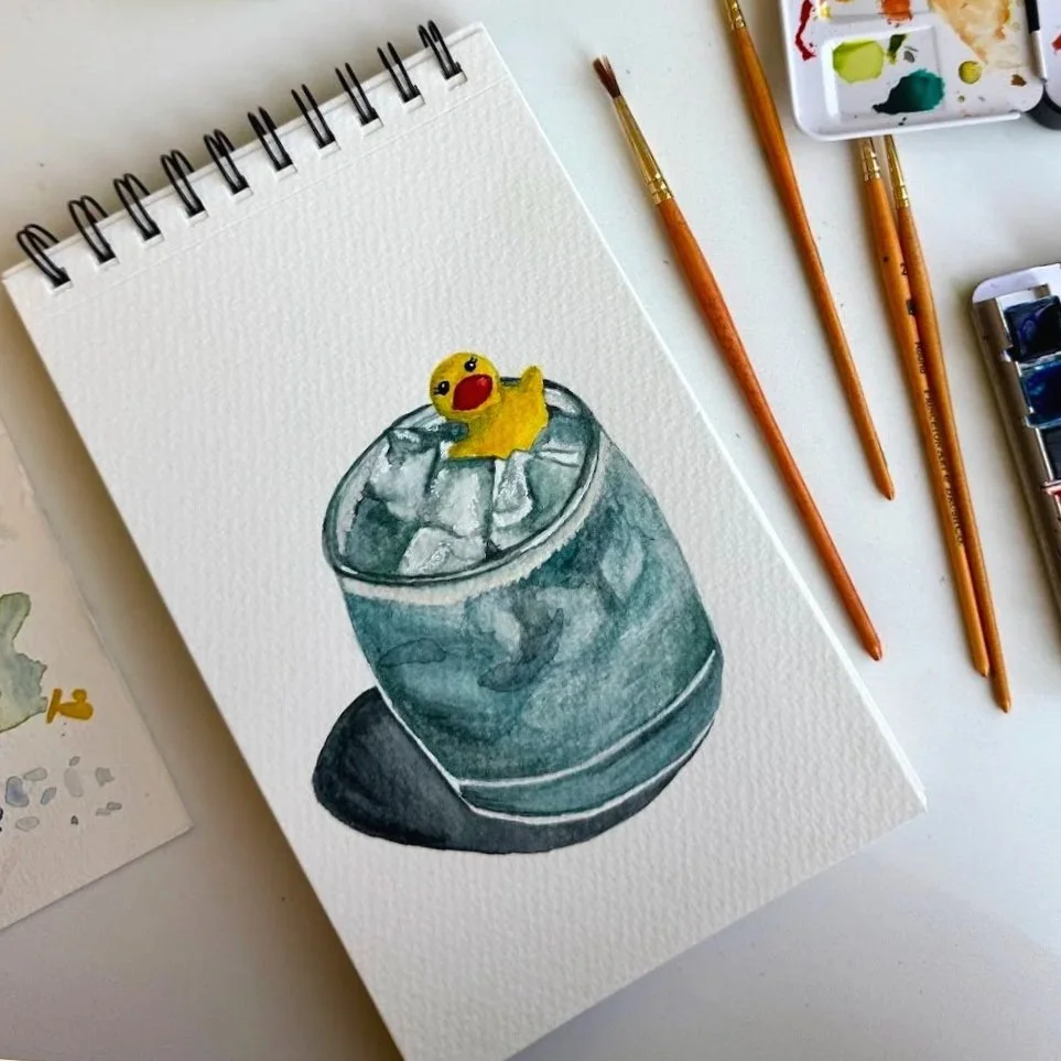 Watercolor painting of a yellow rubber duck in a mocktail on a sketchbook page, with paintbrushes and watercolor palette beside it.