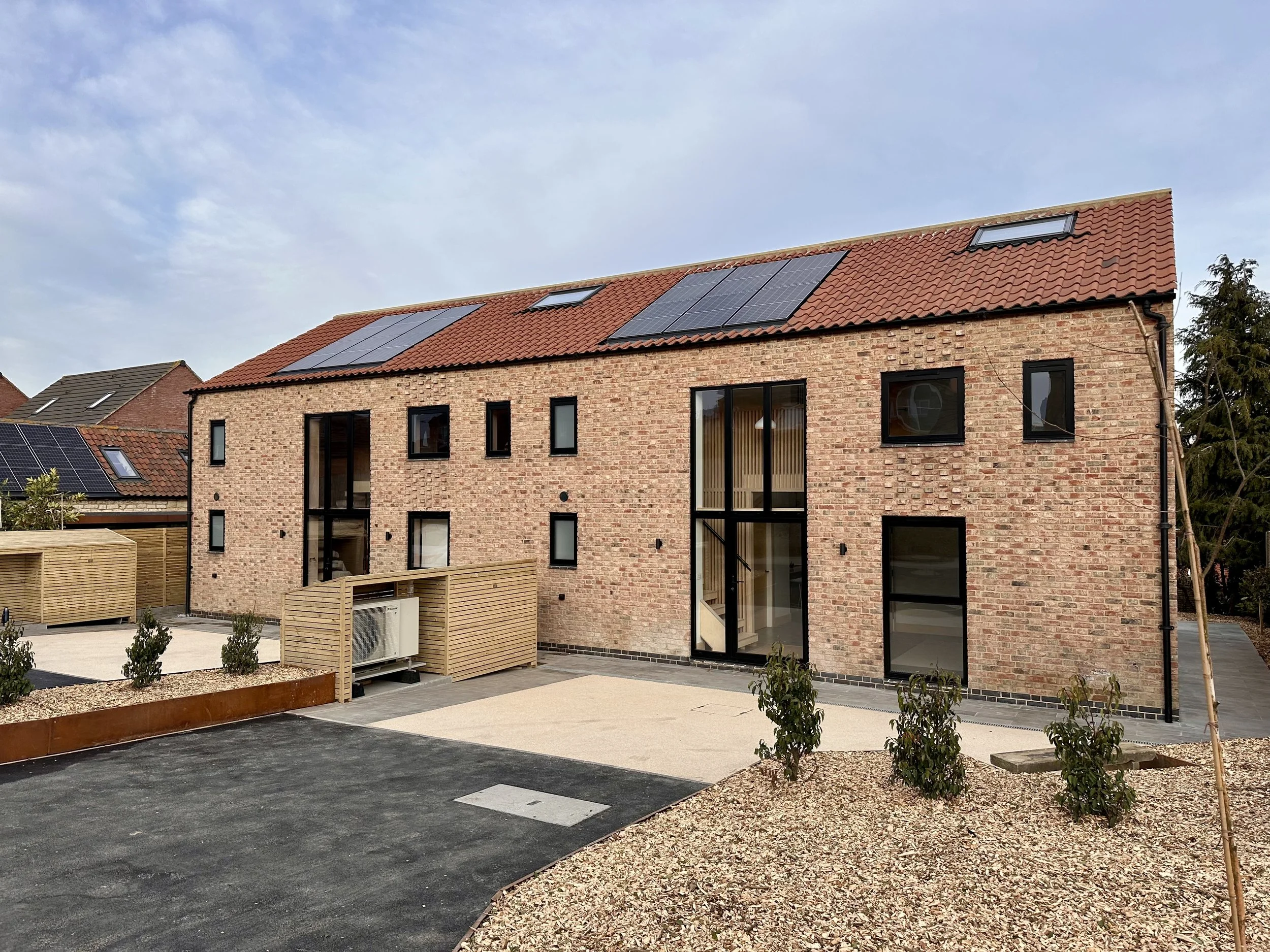 A modern brick residential building with large glass doors and windows, solar panels on the red tiled roof, and landscaped yard with small bushes and gravel.