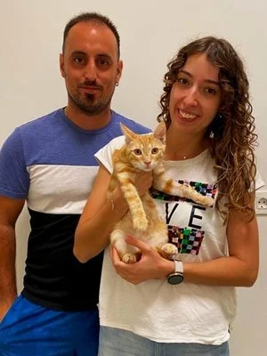Una pareja sostiene un gato naranja en una habitación con fondo claro.