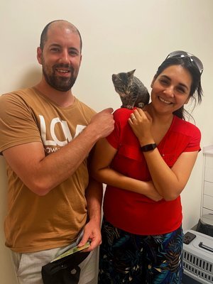 Un hombre y una mujer sonriendo, con un gato en la mano de la mujer, en un ambiente interior.