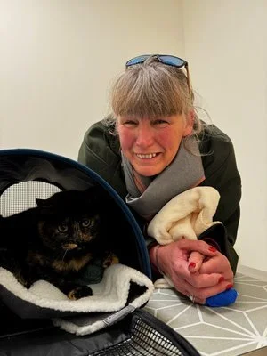 Una joven sonriente con gafas en la cabeza, apoyada en una mesa, con un gato en una mochila y una cobija en las manos.