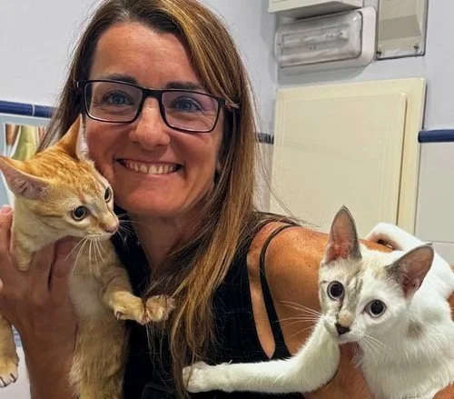 Una mujer con gafas sosteniendo dos gatos, uno naranja y uno blanco, en un entorno interior.