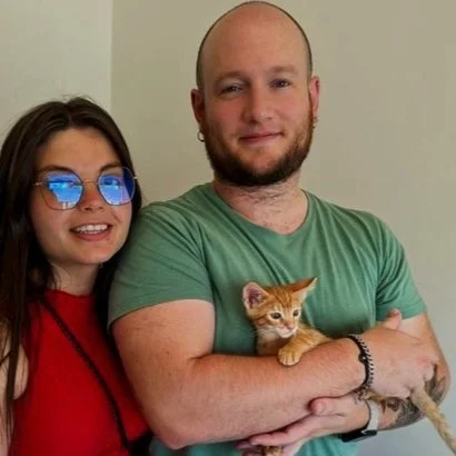 Un hombre y una mujer posando juntos, él sostiene un gato naranja en sus brazos, todos en un entorno interior.