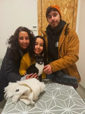 Dos adultos y una niña sonriendo, sosteniendo un gato negro y blanco en una mesa, con una mujer usando un abrigo negro y el hombre usando una chaqueta amarilla. Un fondo de pared de madera y una mesa con un patrón geométrico.