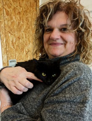 Una mujer con cabello rizado sosteniendo un gato negro en brazos, sonriendo frente a un fondo de madera.
