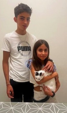 Un joven y una niña sosteniendo un gato blanco con negro, posando frente a una pared blanca con diseño en la parte inferior.