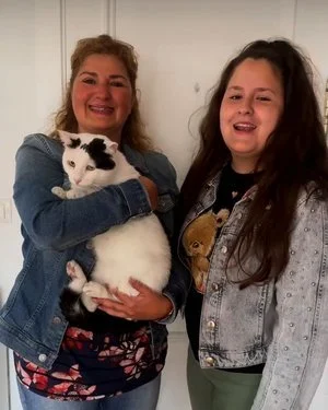 Dos mujeres sonriendo y sosteniendo un gato blanco con manchas negras en un fondo blanco.
