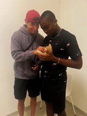 Dos muchachos, uno con gorra roja y sudadera gris, y otro con cabello rapado y camiseta negra, miran y sostienen un objeto en las manos, en un ambiente interior.