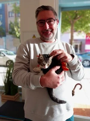 Hombre sonriente sosteniendo un gato en un interior con ventanas y árboles visibles al fondo.