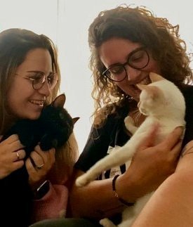 Dos mujeres sonriendo y sosteniendo gatos, uno negro y otro blanco, en un ambiente acogedor.