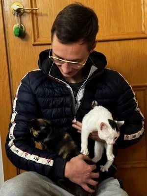 Un joven sosteniendo dos gatos, uno negro con naranja y uno blanco, en un ambiente interior con paneles de madera.